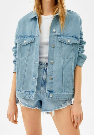 Kobieta w jasnoniebieskiej oversize kurtce dżinsowej na białym prążkowanym topie oraz wysokich, postrzępionych szortach dżinsowych, stojąca na białym tle.