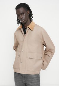 Veste beige avec col en velours côtelé, boutons-pression et deux poches avant, confectionnée dans un tissu lisse et résistant. Coupe décontractée au design moderne.
