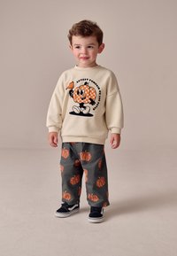 Sweat-shirt crème avec un motif citrouille, associé à un pantalon gris décoré de motifs de citrouilles orange. Des chaussures noires complètent la tenue.