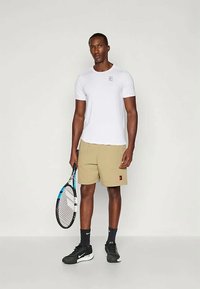 Homem em pé a segurar uma raquete de ténis, vestindo uma t-shirt branca, calções bege, meias pretas e ténis Nike pretos contra um fundo simples.