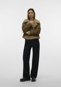 Giacca in shearling marrone con colletto ampio e chiusura con zip, abbinata a jeans a gamba larga blu scuro e stivali alla caviglia marroni. Design semplice.