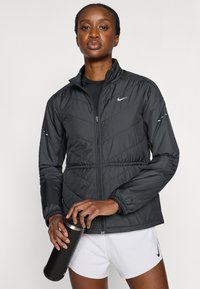 Μαύρο, φουσκωτό μπουφάν Nike με ψηλό γιακά, φερμουάρ μπροστά και ελαστικά μανίκια. Διαθέτει ραφές σε σχήμα βέλους και ανακλαστικές λεπτομέρειες στα μανίκια.