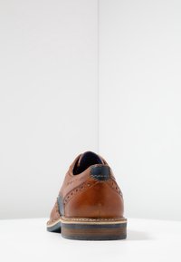 Scarpa in pelle marrone con dettagli brogue, un accento blu navy sul tallone e una suola stratificata con sezioni in legno e gomma.