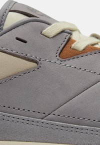 Gros plan sur une chaussure de sport en daim gris avec des accents beige et marron et des lacets de couleur crème passés à travers des œillets.