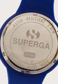 Sinine silikoonkell, millel on roostevabast terasest tagaosa, millel on graveeritud "SUPERGA," "VEELÄBIVUS 3 ATM" ja "STC084." Sujuv tekstuur.