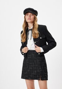 Sort tweed blazer med sølvknapper, hvid flæserbluse og sort nederdel med sideslids. Sort kasket med kædeaccent.