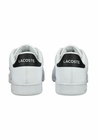 Bílé kožené tenisky s černým patním tabem, s logem "LACOSTE". Hladký povrch, minimalistický design a šněrování.