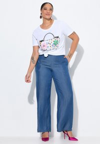 Biała koszulka z grafiką torby z kwiatami, niebieskie spodnie dzwony i różowe buty na wysokim obcasie. Modelka pozuje z rękami na biodrach i związanym T-shircie.