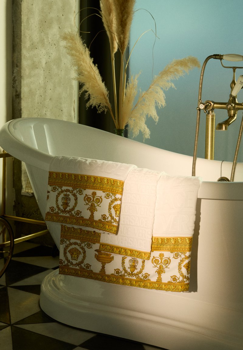 Des serviettes blanches avec des motifs dorés ornés et le texte "VERSACE" en relief pendent sur le rebord d'une baignoire autoportante blanche dans une salle de bains élégante.