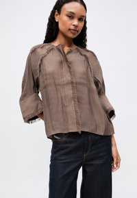 Frau in lockerer brauner Bluse mit ausgefransten Kanten und dunkelblauer Jeans, die vor einem schlichten hellen Hintergrund steht.