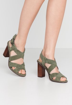 Femme portant des sandales à talons hauts en daim vert avec des talons blocs en bois et des brides croisées sur un sol gris clair.