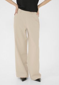 Pantalons large beiges en tissu lisse, dot és d'une taille haute et de poches latérales, associés à des escarpins noirs pointus.