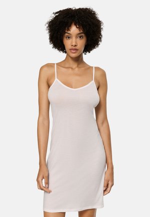 Ung kvinde med krøllet hår iført en lyserød, ærmeløs slipkjole med tynde stropper, stående foran en ensfarvet baggrund.