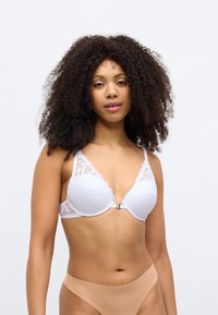 Valkoinen pitsirintaliivit, joissa on aaltoilevat reunat, sileä kangaskeskiö ja metallinen kiinnike. Yhdistetty beigeisiin korkeavyötäröisiin alushousuihin.