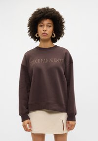 Sweatshirt castanho com "DOLCE FAR NIENTE" bordado em tom beje, com um corte relaxado e ombros caídos, combinado com uma saia bege clara.