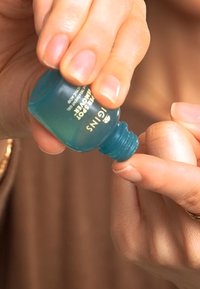 Turkis glassflaske med hvit og gull tekst, holdt i en vinkel. Væske blir dispenset på en fingertupp. Glatt tekstur.