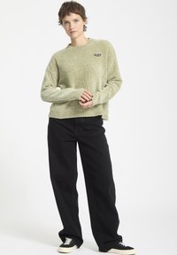 Pull en maille verte avec un col rond et un détail en patch noir, associé à un pantalon large noir et des chaussures noires avec des semelles blanches.