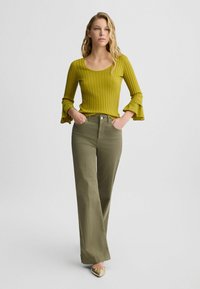 Zelený pletený top s dlouhými rukávy a rozšířenými manžetami, kombinovaný s olivovými kalhotami širokého střihu. Modelka má na sobě zlaté baleríny s mašlí.