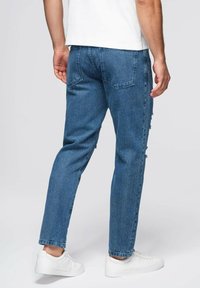 Jeans de mezclilla azul con un corte holgado, costuras visibles, dos bolsillos traseros y detalles desgastados cerca del dobladillo, combinados con zapatillas blancas.