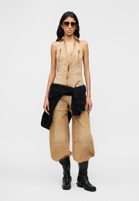 Femme portant un haut sans manches couleur beige avec fermeture éclair, un pantalon beige ample rentré dans des bottes noires, une veste noire nouée à la taille, et des lunettes de soleil.