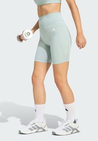 Svetlozelené športové šortky s vysokým pásom, so bočnými vreckami a logom Adidas. Skombinované s bielymi topánkami a činkou.