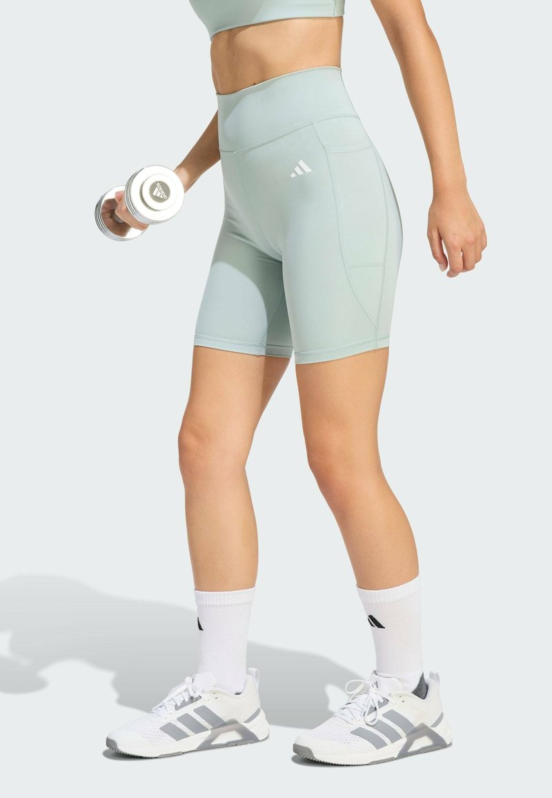 Svetlozelené športové šortky s vysokým pásom, so bočnými vreckami a logom Adidas. Skombinované s bielymi topánkami a činkou.