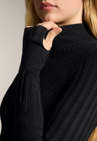 Žena u crnom džemperu rebrastog pletenja s rupom za palac, dodirujući ovratnik, pokazujući dio lica i plavu kosu.