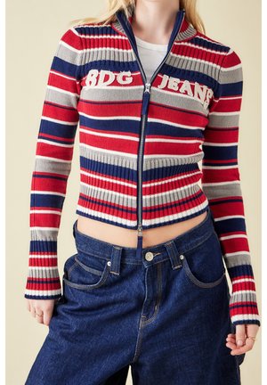 Kvinde iført en rød, marineblå, grå og hvidstribet lynlåstrøje med teksten "BDG JEANS" og højttaljede mørkeblå jeans.