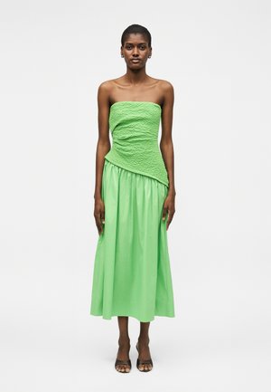 Femme debout portant une robe verte bustier avec un haut texturé et une jupe fluide, associée à des talons ouverts noirs sur fond blanc.