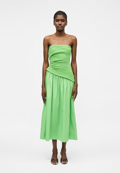 Femme debout portant une robe verte bustier avec un haut texturé et une jupe fluide, associée à des talons ouverts noirs sur fond blanc.
