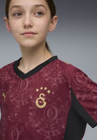 Punainen ja musta urheilupaita, jossa on V-kaula-aukko, kultaiset yksityiskohdat, logo ja etuosassa kukkakuosi.