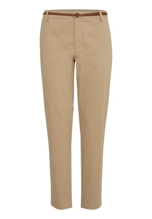 Frau trägt beige Slim-Fit-Hose, weißes T-Shirt mit halbem Arm und weiße Sneakers, steht mit den Händen in den Taschen vor einem einfarbigen Hintergrund.