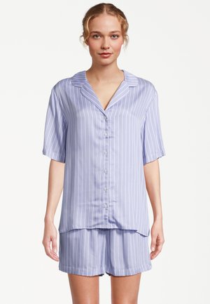 Kobieta ubrana w jasnoniebieską koszulę na krótki rękaw z pionowymi białymi paskami oraz dopasowane szorty, stojąca na białym tle.