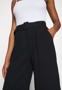 Svarta, högmidjade byxor med vida ben, utrustade med ett bälte med fyrkantig spänne, veck framtill och sidofickor. Texturerat tyg.