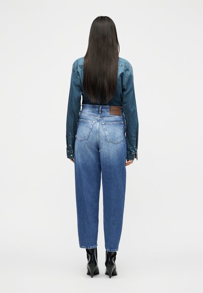 Γυναίκα με μακριά ίσια μαλλιά, ντυμένη με σκουρόχρωμο τζιν πουκάμισο, ανοιχτόχρωμο τζιν ψηλόμεσο παντελόνι και μαύρα μποτάκια με τακούνι, στέκεται με την πλάτη γυρισμένη.