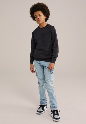 Czarny sweatshirt z kieszenią na piersi, zestawiony z jasnoniebieskimi dżinsami typu cargo z bocznymi kieszeniami. Całość dopełniają czarno-białe sneakersy.