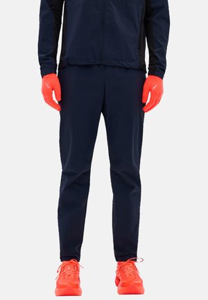 Person wearing black pants, black jacket, bright orange gloves, and bright orange running shoes standing on a plain white background.