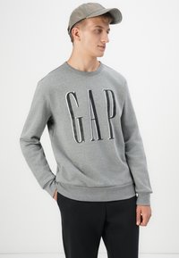 Sudadera gris con gran logo "GAP" en negro. Corte regular, cuello redondo, mangas largas, puños y dobladillo acanalados. Combinada con pantalones negros.