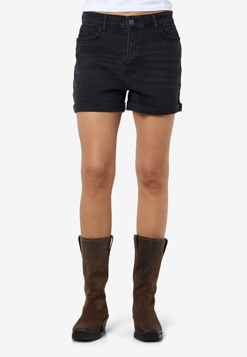 Personne portant un short en jean noir à ourlet roulé et des bottes en cuir marron montant jusqu'au milieu du mollet, debout devant un fond blanc uni.