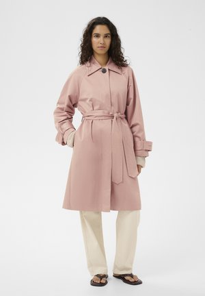 Vrouw met krullend haar die een roze trenchcoat met ceintuur, beige broek en donkere slippers draagt, staand tegen een effen witte achtergrond.