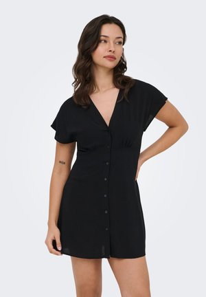 Jeune femme portant une robe noire à manches courtes avec boutons sur le devant, posant avec la main sur la hanche, sur un fond clair uni.