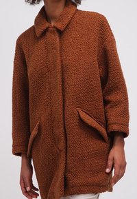 Brun texturerad rock med krage, knappstängning framtill och sidofickor. Tillverkad av ett fluffigt material, med en avslappnad passform.