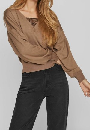Femme portant un pull ample marron avec un caraco en dentelle noire en dessous et un jean noir taille haute sur un fond uni.