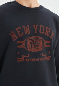 Sudadera de un oscuro tono azul marino con un gráfico rojo prominente que dice "NUEVA YORK," un motivo de fútbol y la frase "UN EQUIPO UN SUEÑO."