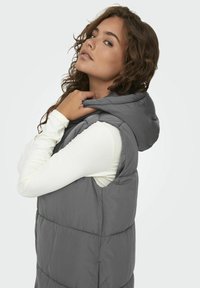 Gilet à capuche gris avec un design matelassé, présentant une texture douce. Porté par-dessus une chemise à manches longues de couleur claire, montrant une coupe décontractée.