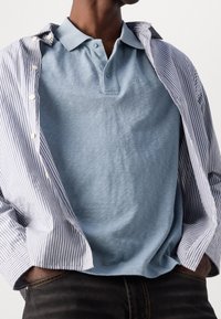 Ljusblå pikétröja med mjuk textur, bärs under en blå och vit randig knappskjorta. Märkbart krage och diskret varumärkesdetalj.