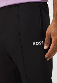 Schwarze Sporthosen aus strukturiertem Stoff, mit einer Seitentasche und einem weißen "BOSS"-Logo auf dem Oberschenkel.
