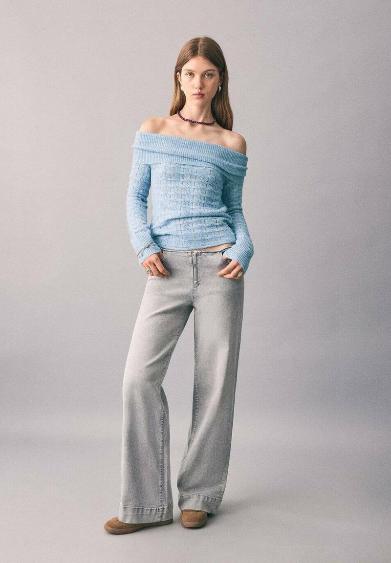 O tânără stă cu mâinile în buzunare purtând un pulover deschis la culoare, de culoare albastru deschis, blugi gri cu picioare largi, pantofi negri și un colier violet.