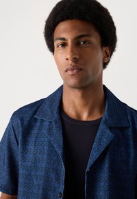 Marineblaues Kurzarmhemd mit einem geometrischen Muster in einem helleren Farbton, ausgestattet mit einem abgeschnittenen Kragen und Knopfleiste. Darunter befindet sich ein schwarzes, gerippte T-Shirt.