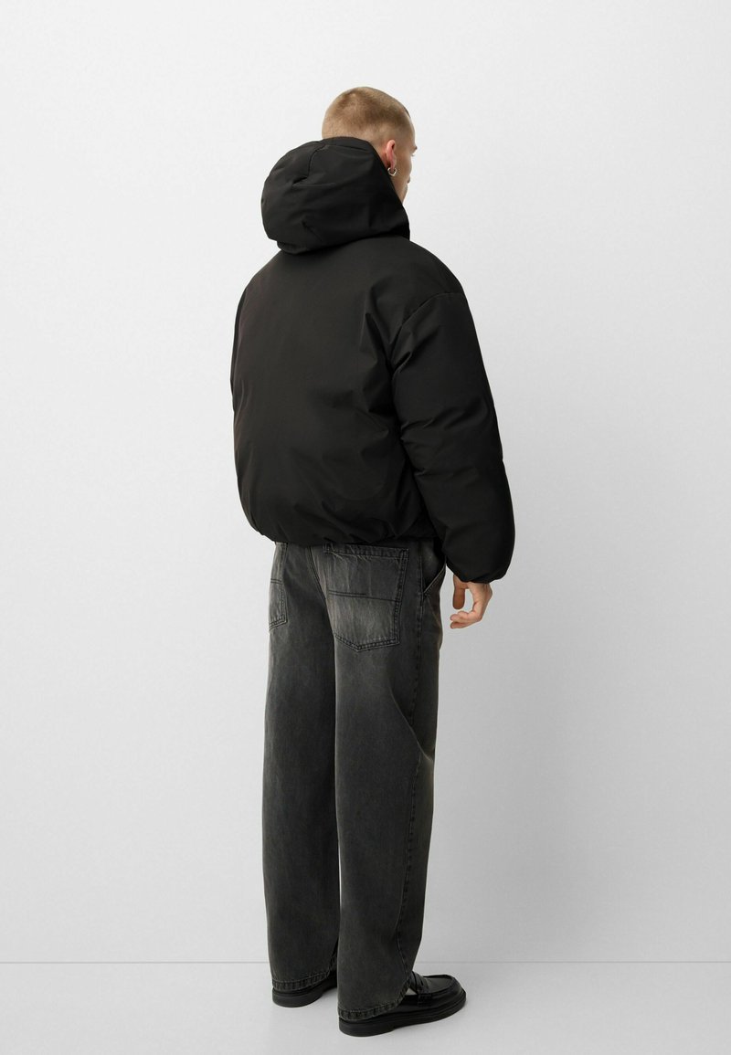 Giubbotto a puffo nero con cappuccio, caratterizzato da una silhouette arrotondata e una texture liscia. Indossato con pantaloni in denim grigi larghi e scarpe nere.